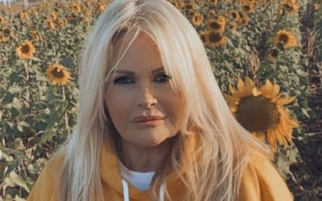 A ex-modelo Monique Evans com moletom laranja, expressão séria, posa para foto em ambiente externo, com sol e plantas ao fundo