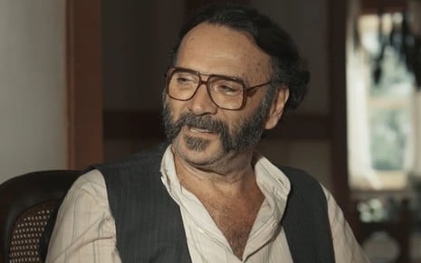 Almir Sater caracterizado como Rachid; ele usa uma barba longa, veste camisa e paletó e dá um leve sorriso em cena de Renascer